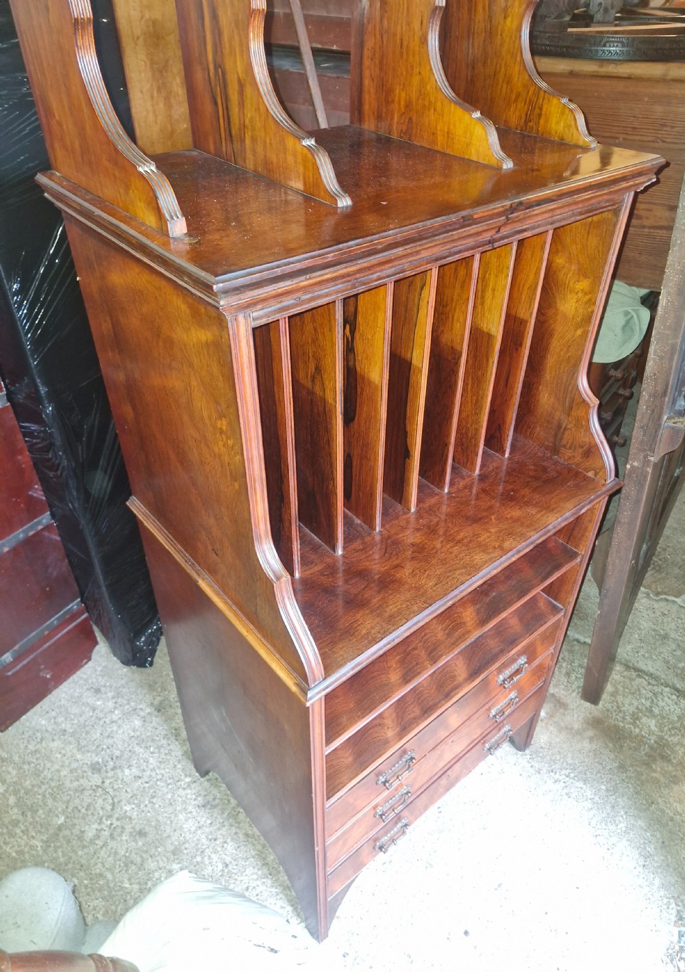 A late Victorian dark rosewood side cabinet featuring upper tiered display shelves, vertical open folio partitions, lower open shelving, and small drawers with brass drop handles. View V
