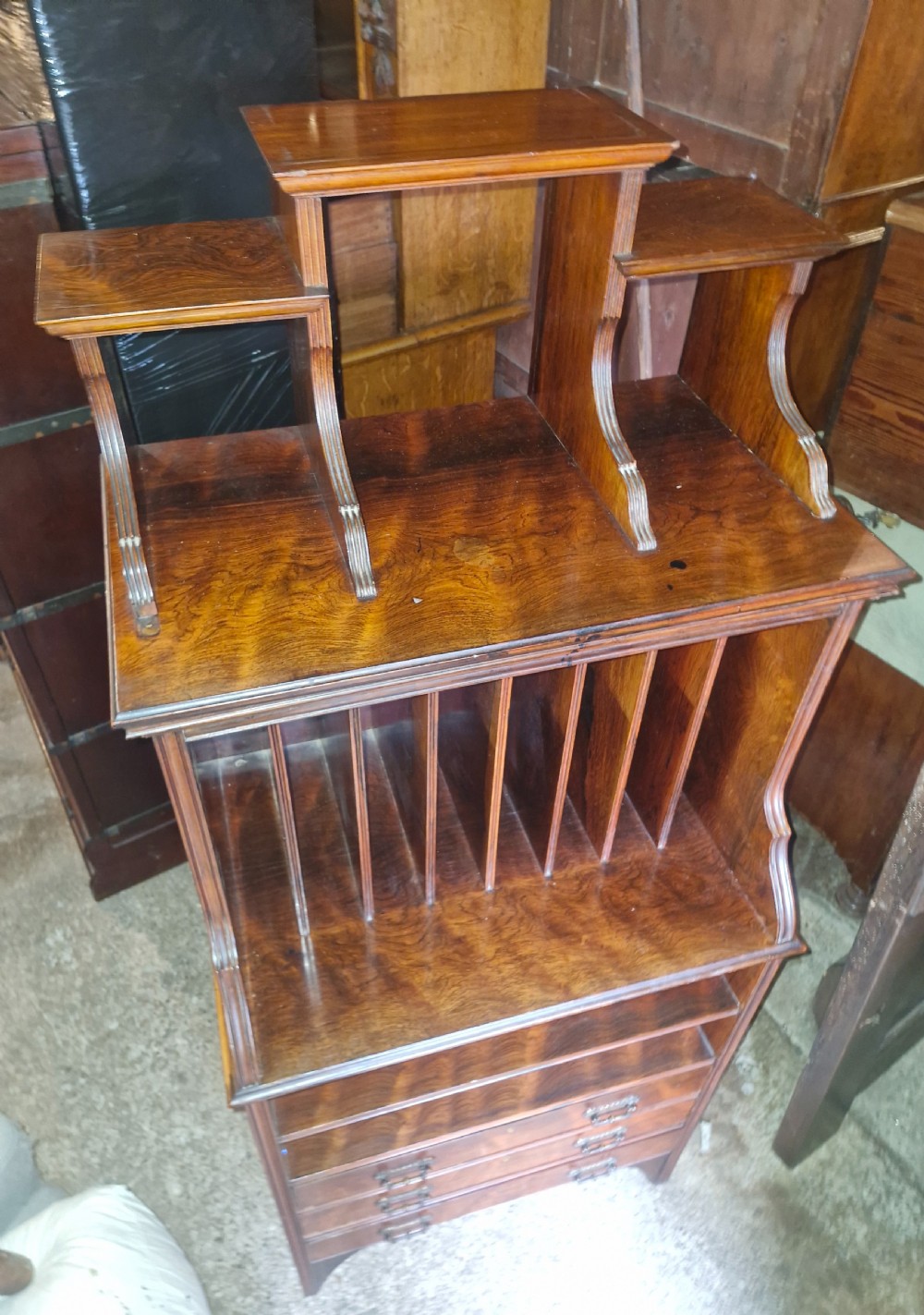 A late Victorian dark rosewood side cabinet featuring upper tiered display shelves, vertical open folio partitions, lower open shelving, and small drawers with brass drop handles. View VII
