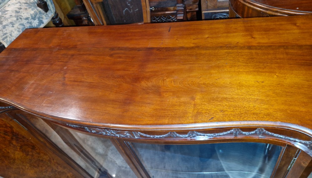 An Edwardian mahogany display cabinet featuring carved floral swags, two glazed doors, glass shelves, and a blue patterned fabric backing. View III