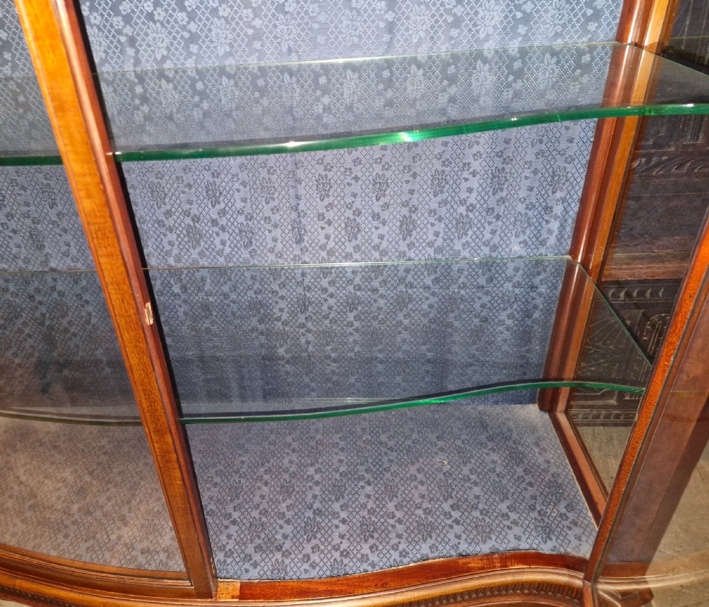 An Edwardian mahogany display cabinet featuring carved floral swags, two glazed doors, glass shelves, and a blue patterned fabric backing. View V