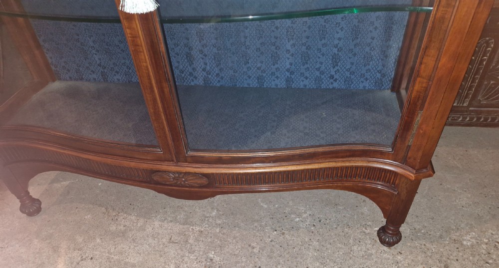 An Edwardian mahogany display cabinet featuring carved floral swags, two glazed doors, glass shelves, and a blue patterned fabric backing. View VI