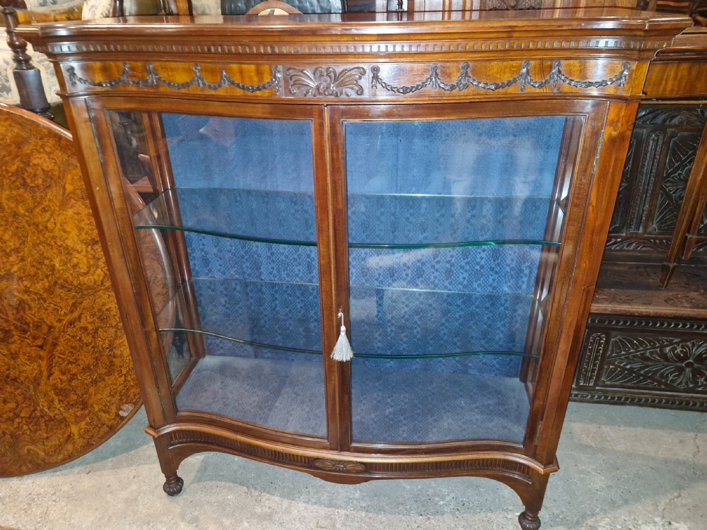 An Edwardian mahogany display cabinet featuring carved floral swags, two glazed doors, glass shelves, and a blue patterned fabric backing. View VII