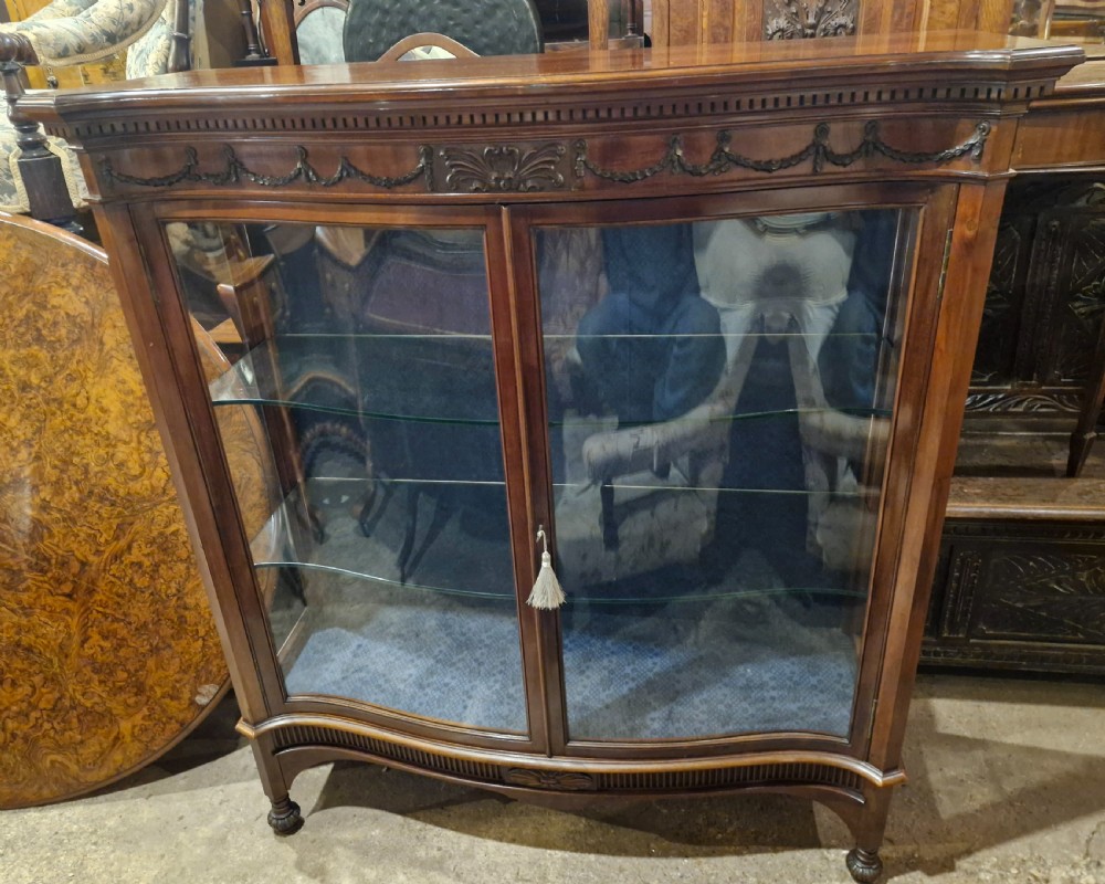 An Edwardian mahogany display cabinet featuring carved floral swags, two glazed doors, glass shelves, and a blue patterned fabric backing. View X