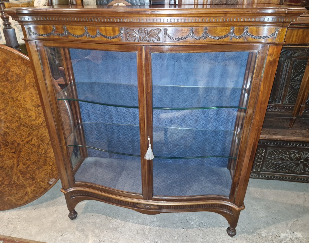 An Edwardian mahogany display cabinet featuring carved floral swags, two glazed doors, glass shelves, and a blue patterned fabric backing.