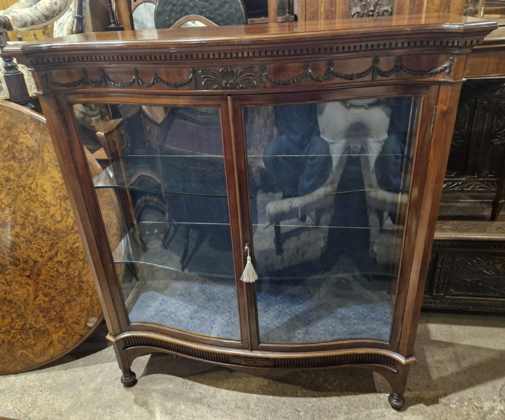 An Edwardian mahogany display cabinet featuring carved floral swags, two glazed doors, glass shelves, and a blue patterned fabric backing. View II
