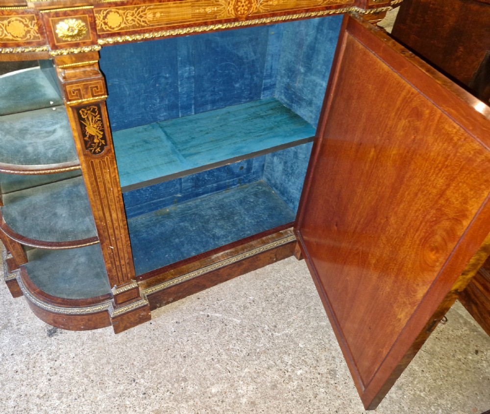 An antique burr walnut side cabinet featuring intricate marquetry inlays, gilt bronze mounts, a central mirror, and open curved side shelves lined in green velvet. View VI