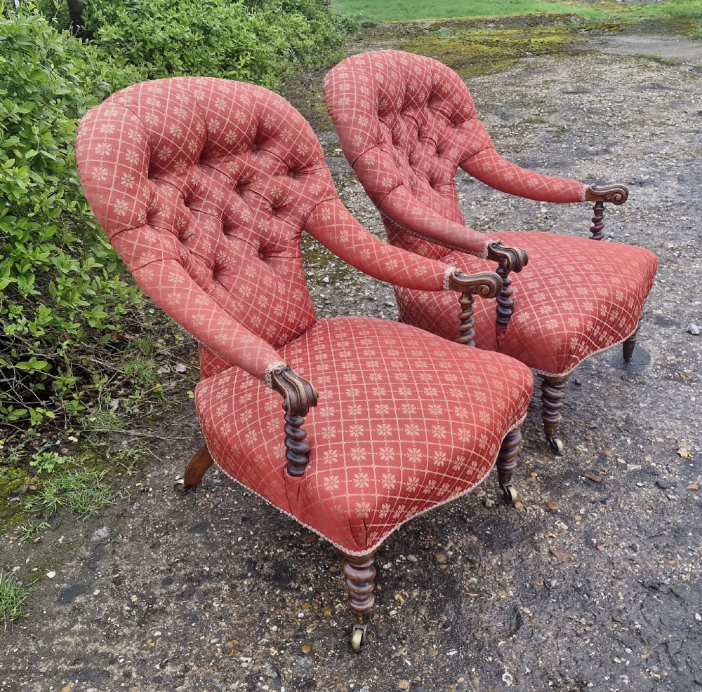 A pair of Victorian armchairs upholstered in patterned terracotta red fabric featuring button-tufted backs, barley twist arm supports, and carved wooden legs on brass castors.
