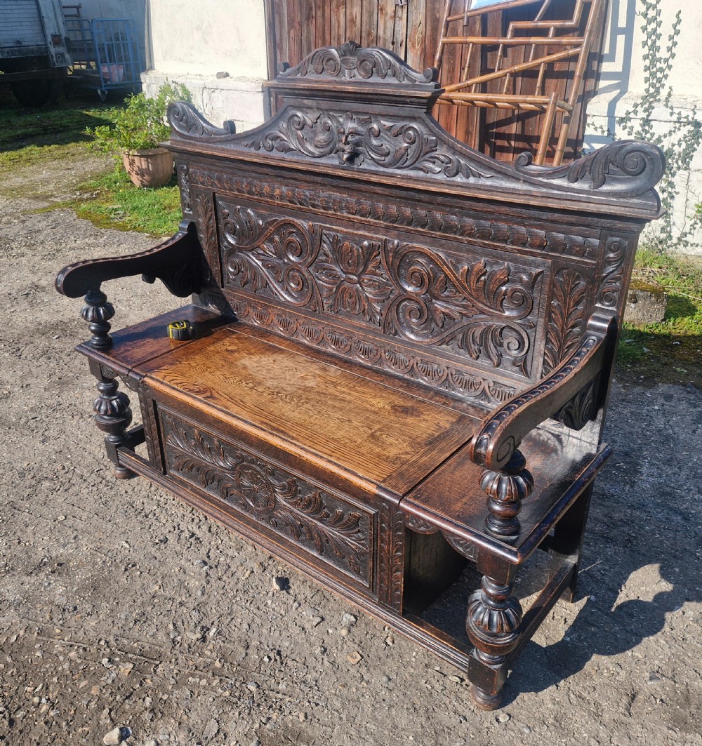 A late Victorian dark carved oak hall bench featuring foliate scrollwork, a central carved mask crest, turned front legs, and a lift-up seat storage compartment. View II