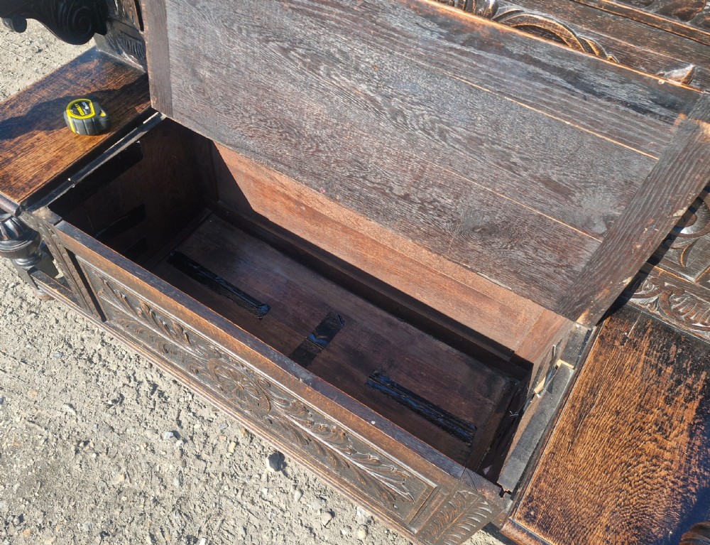 A late Victorian dark carved oak hall bench featuring foliate scrollwork, a central carved mask crest, turned front legs, and a lift-up seat storage compartment. View III