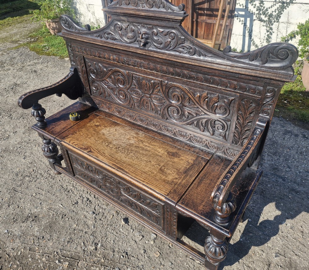 A late Victorian dark carved oak hall bench featuring foliate scrollwork, a central carved mask crest, turned front legs, and a lift-up seat storage compartment. View V