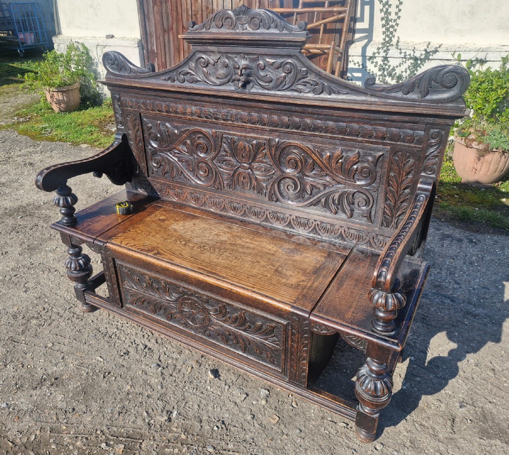 A late Victorian dark carved oak hall bench featuring foliate scrollwork, a central carved mask crest, turned front legs, and a lift-up seat storage compartment. View VI