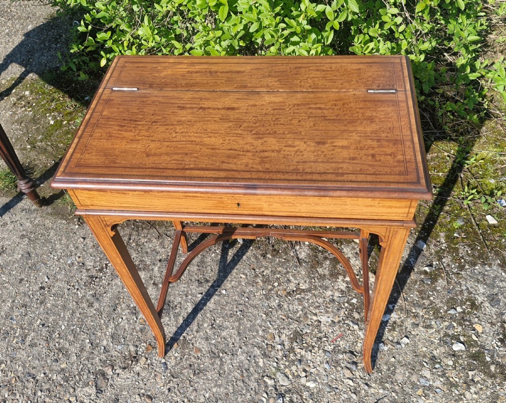 An Edwardian inlaid satinwood writing table featuring a hinged top surface, slender tapered legs with a curved cross-stretcher, and fine dark wood stringing. View XI