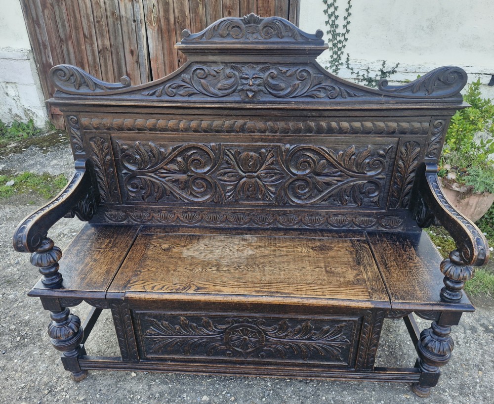 A late Victorian dark carved oak hall bench featuring foliate scrollwork, a central carved mask crest, turned front legs, and a lift-up seat storage compartment.