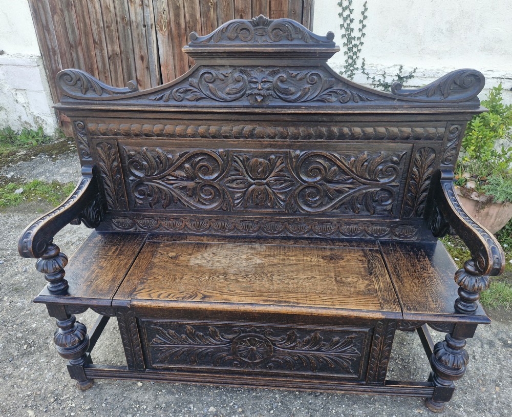 A late Victorian dark carved oak hall bench featuring foliate scrollwork, a central carved mask crest, turned front legs, and a lift-up seat storage compartment. View VII