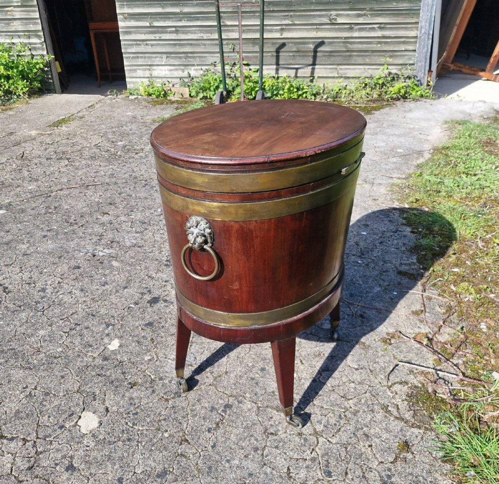 An early nineteenth-century oval mahogany wine cellarette featuring brass binding, lion mask side handles, and tapered legs on brass castors. View II