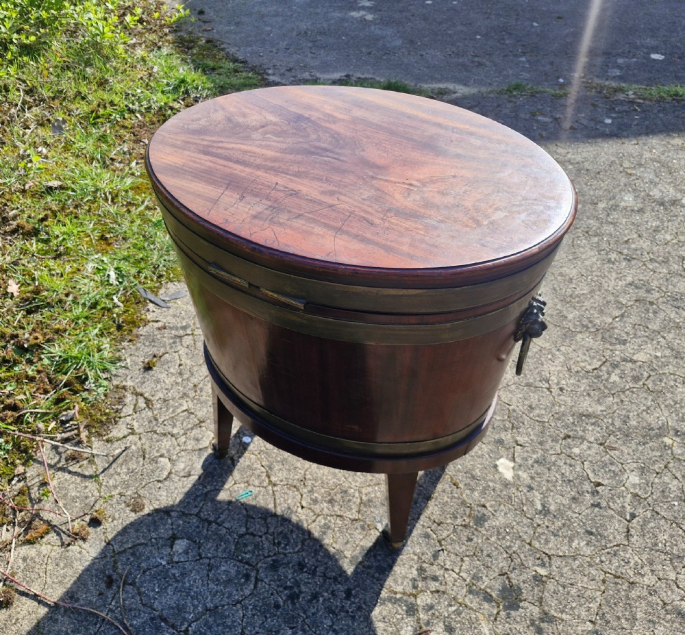 An early nineteenth-century oval mahogany wine cellarette featuring brass binding, lion mask side handles, and tapered legs on brass castors. View VI