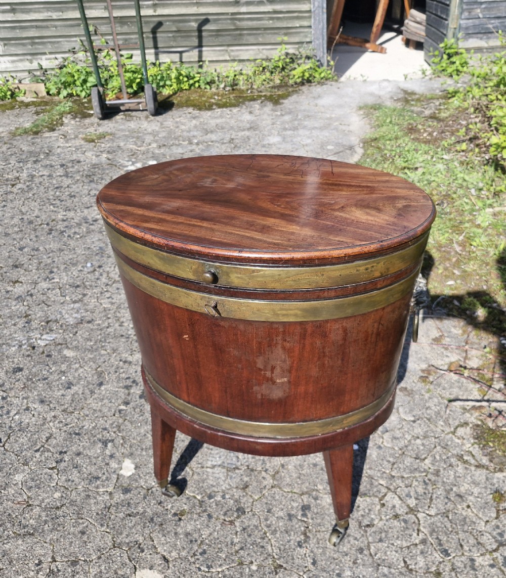 An early nineteenth-century oval mahogany wine cellarette featuring brass binding, lion mask side handles, and tapered legs on brass castors. View X