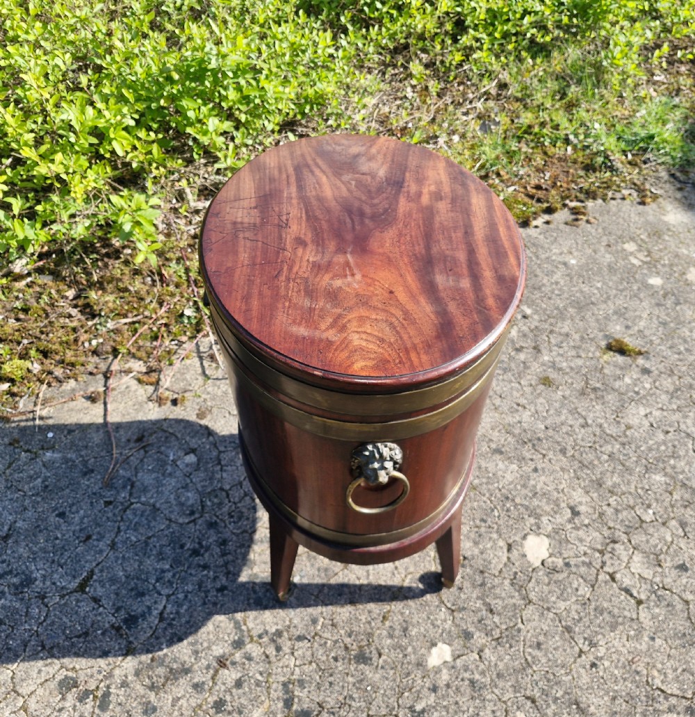 An early nineteenth-century oval mahogany wine cellarette featuring brass binding, lion mask side handles, and tapered legs on brass castors. View XI