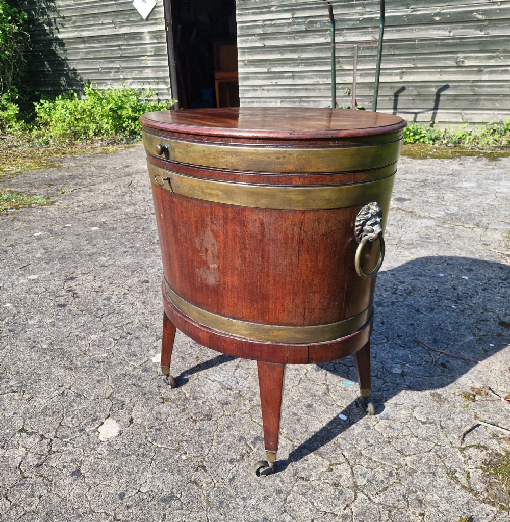 An early nineteenth-century oval mahogany wine cellarette featuring brass binding, lion mask side handles, and tapered legs on brass castors. View XIII