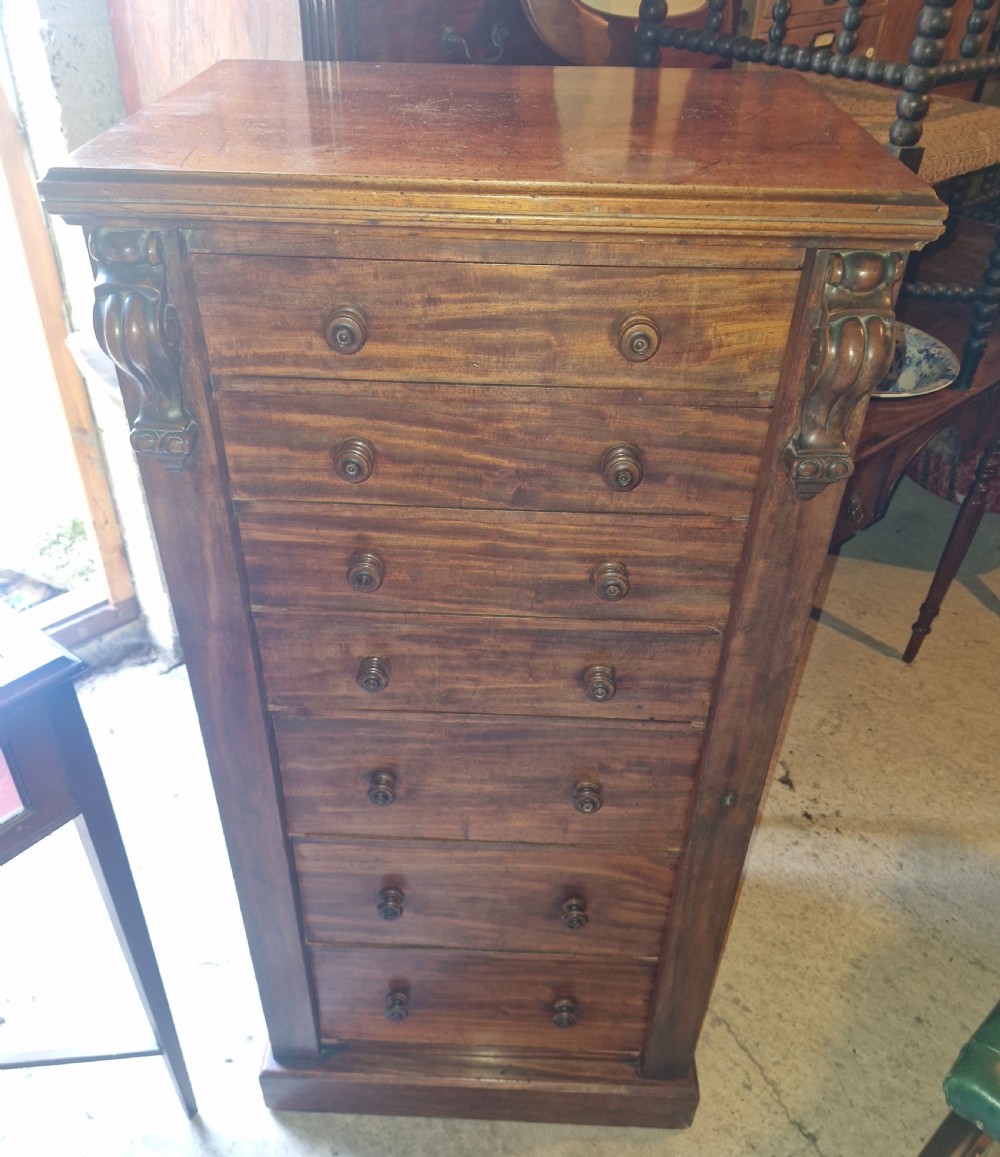 A mid-19th century mahogany Wellington chest featuring seven drawers, wooden knob handles, carved upper corbels, and a right-hand locking bar. View VI