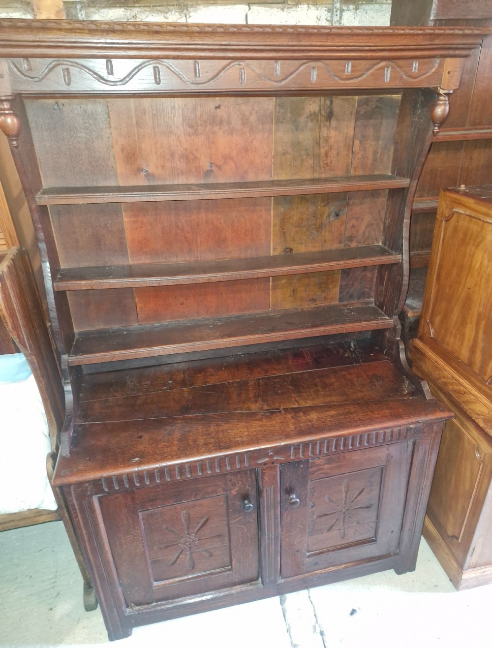 A dark oak cottage dresser featuring a three-tier open plate rack mounted above a two-door base cabinet with carved panel motifs. View VII