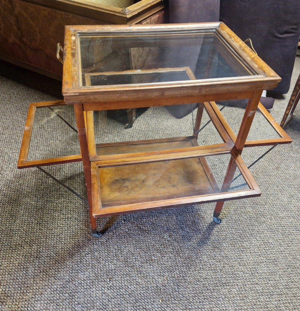 A glazed wooden tea trolley featuring glass side panels, a glass top surface, brass handles, a lower shelf, and castors. View II