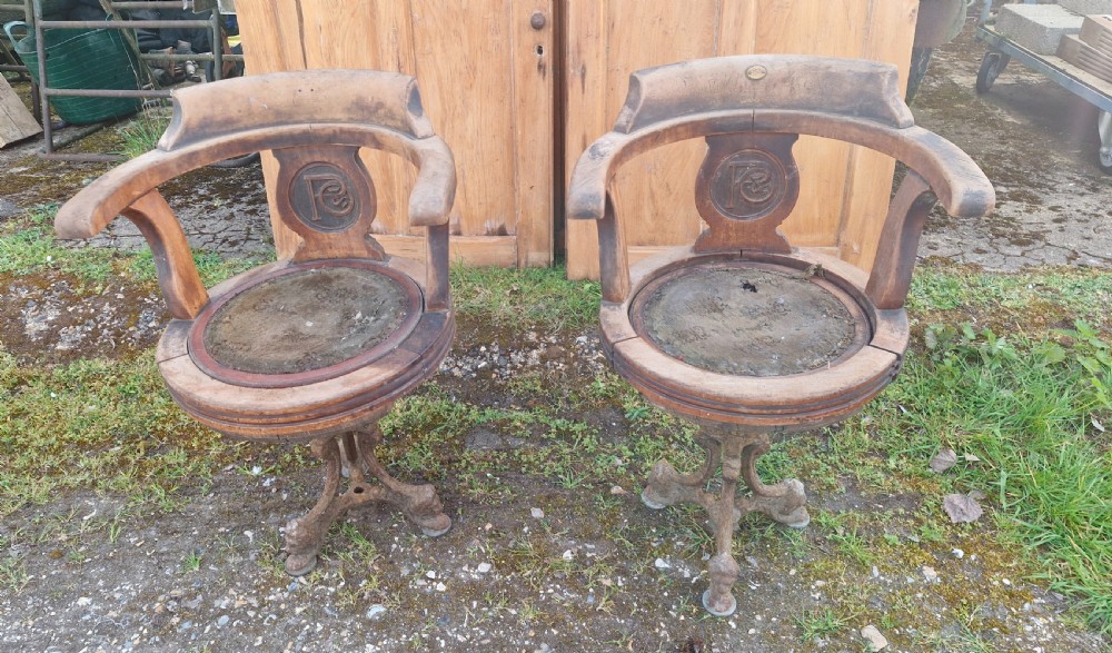 A pair of antique wooden ocean liner revolving armchairs featuring carved monograms on the back splats and cast iron swivel bases.