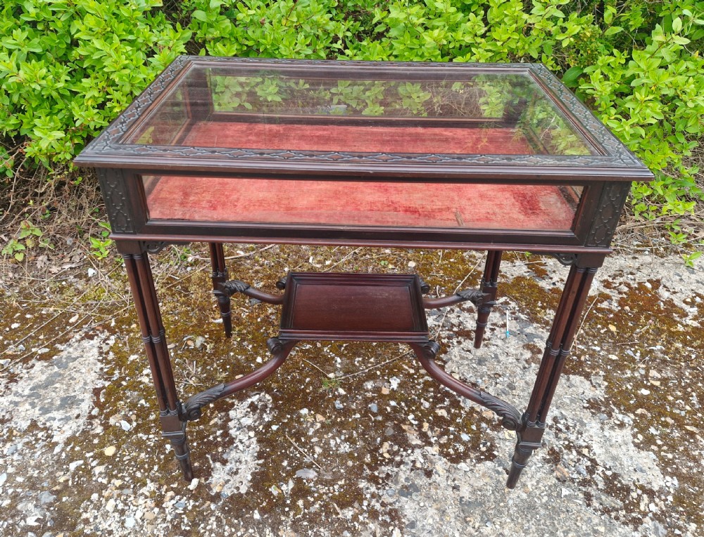 An Edwardian mahogany bijouterie display table featuring a glazed top with red fabric lining, carved fretwork border, and an ornate lower undertier.
