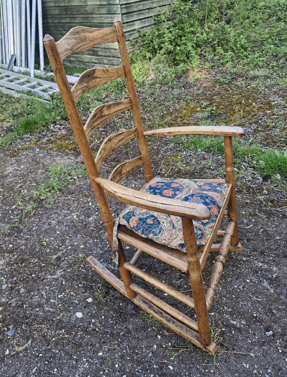 An early 19th century wooden ladderback rocking armchair with curved armrests, turned stretchers, and a patterned cushion.