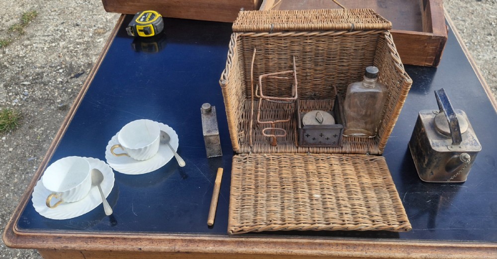 An antique woven wicker travelling hamper displayed open alongside white porcelain teacups, saucers, spoons, a metal spirit kettle, and a glass bottle.