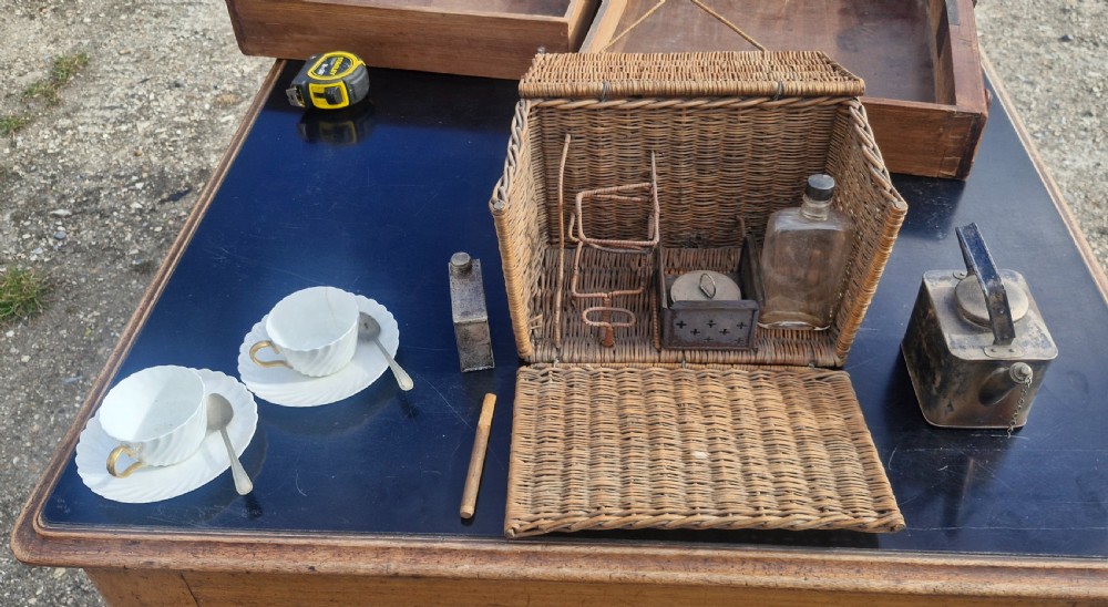 An antique woven wicker travelling hamper displayed open alongside white porcelain teacups, saucers, spoons, a metal spirit kettle, and a glass bottle. View II