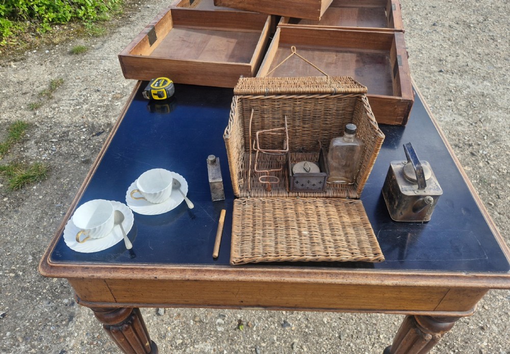 An antique woven wicker travelling hamper displayed open alongside white porcelain teacups, saucers, spoons, a metal spirit kettle, and a glass bottle. View III