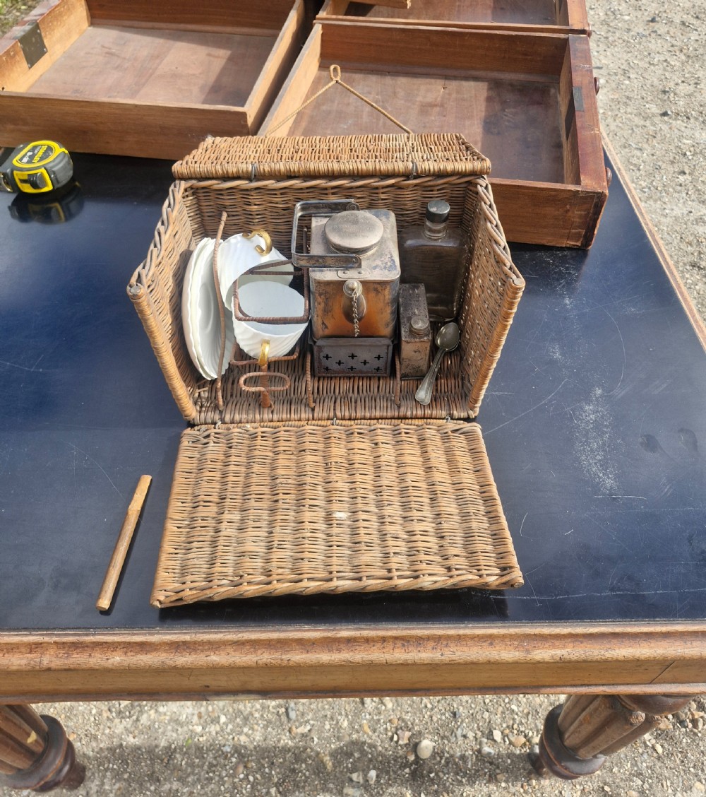 An antique woven wicker travelling hamper displayed open alongside white porcelain teacups, saucers, spoons, a metal spirit kettle, and a glass bottle. View IV