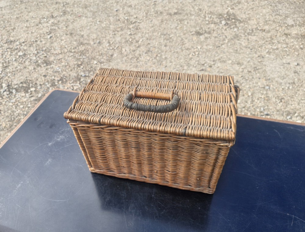An antique woven wicker travelling hamper displayed open alongside white porcelain teacups, saucers, spoons, a metal spirit kettle, and a glass bottle. View V