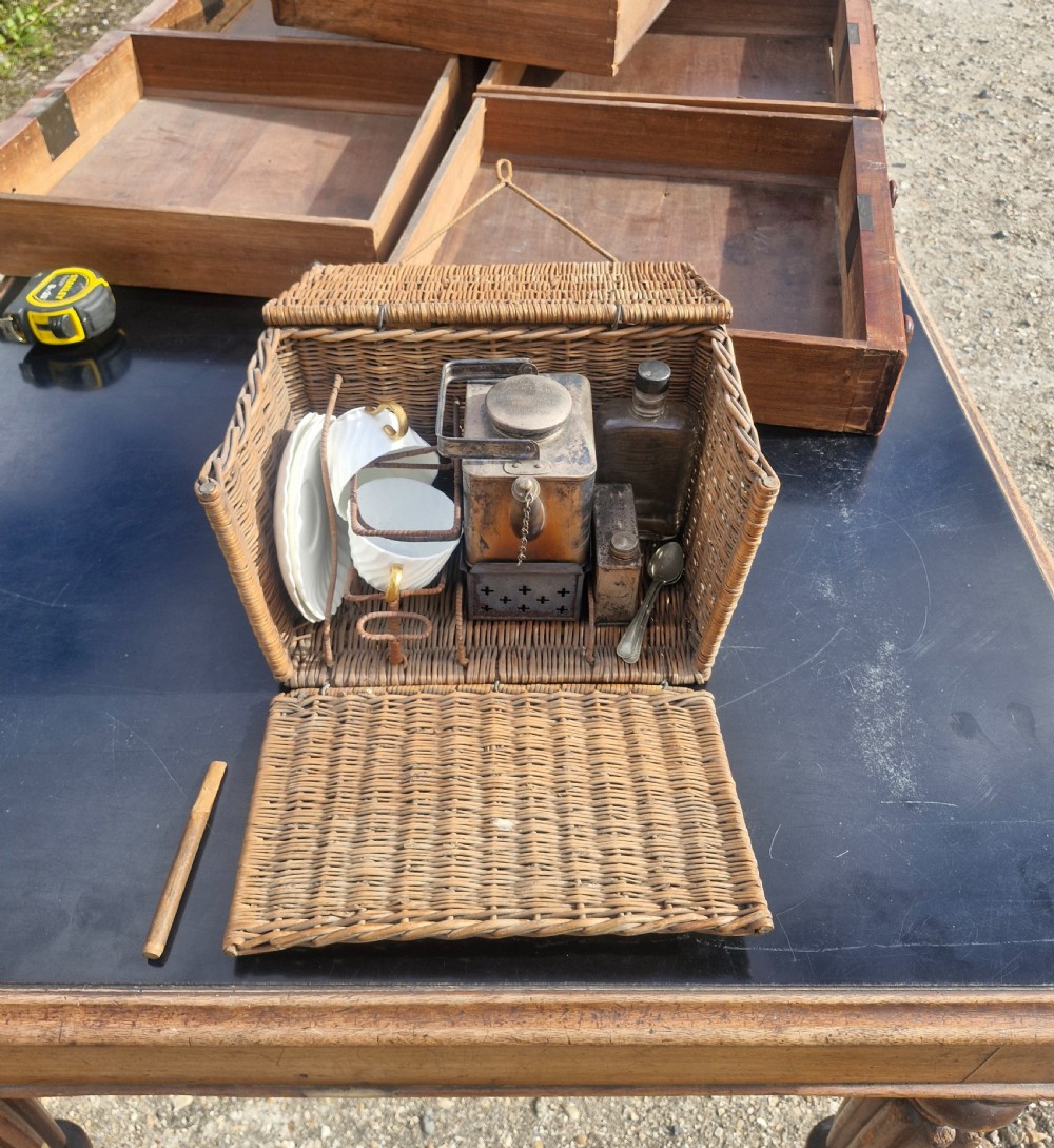 An antique woven wicker travelling hamper displayed open alongside white porcelain teacups, saucers, spoons, a metal spirit kettle, and a glass bottle. View VI