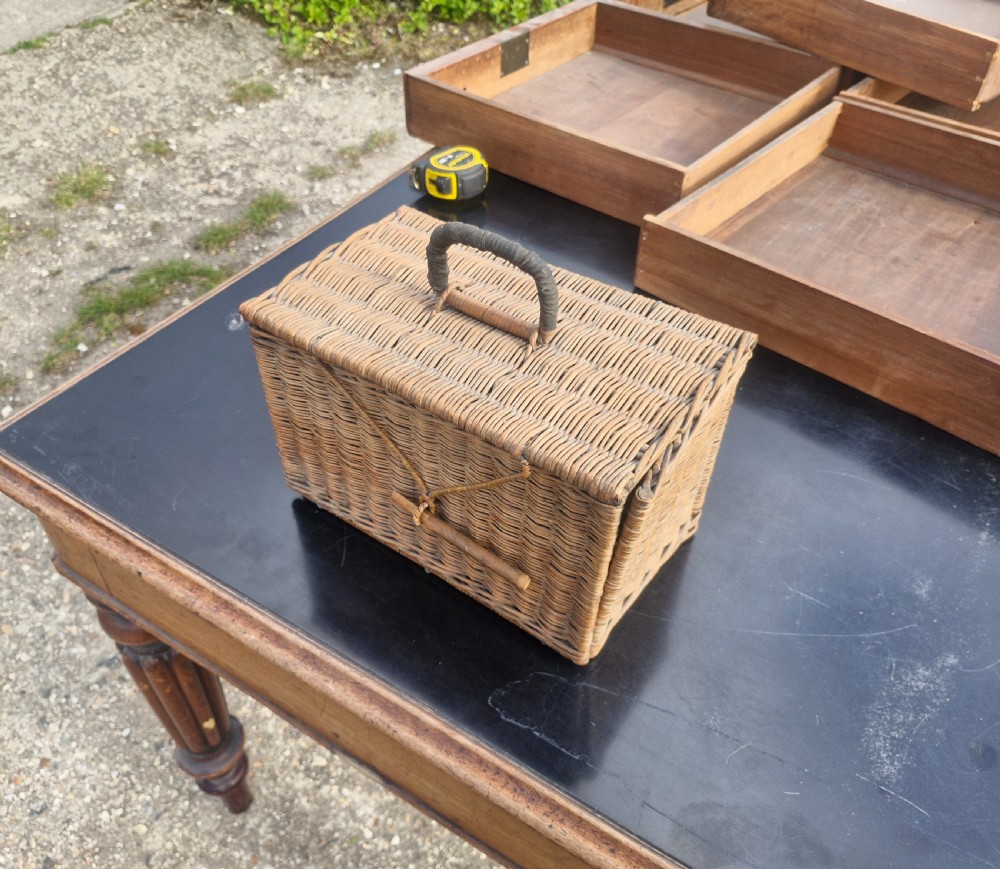 An antique woven wicker travelling hamper displayed open alongside white porcelain teacups, saucers, spoons, a metal spirit kettle, and a glass bottle. View VII