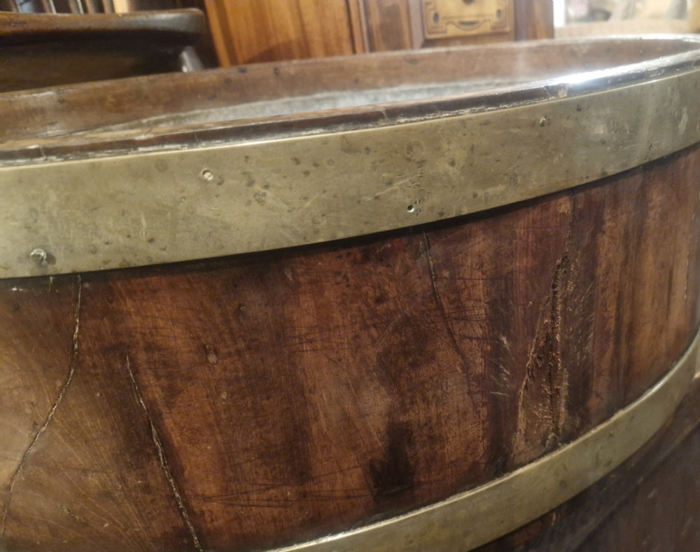 An oval late Georgian mahogany wine cooler with dual brass bands, brass lion mask ring handles, resting on a four-legged stand. View IX