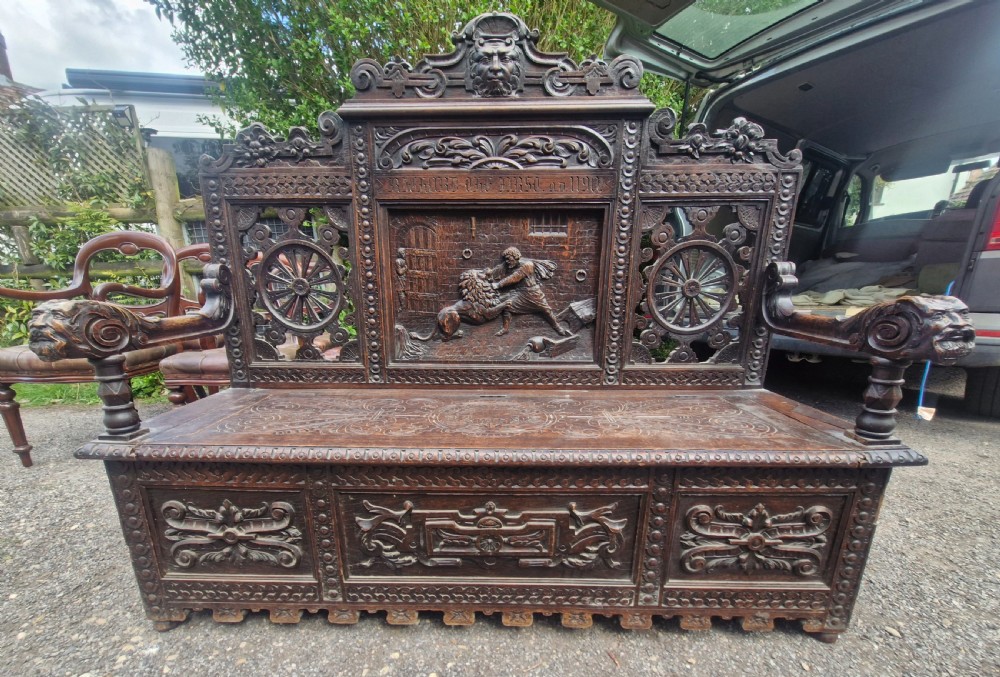 A late nineteenth-century dark carved oak hall bench featuring relief narrative carving, mask pediment, scroll arms, and a hinged seat.