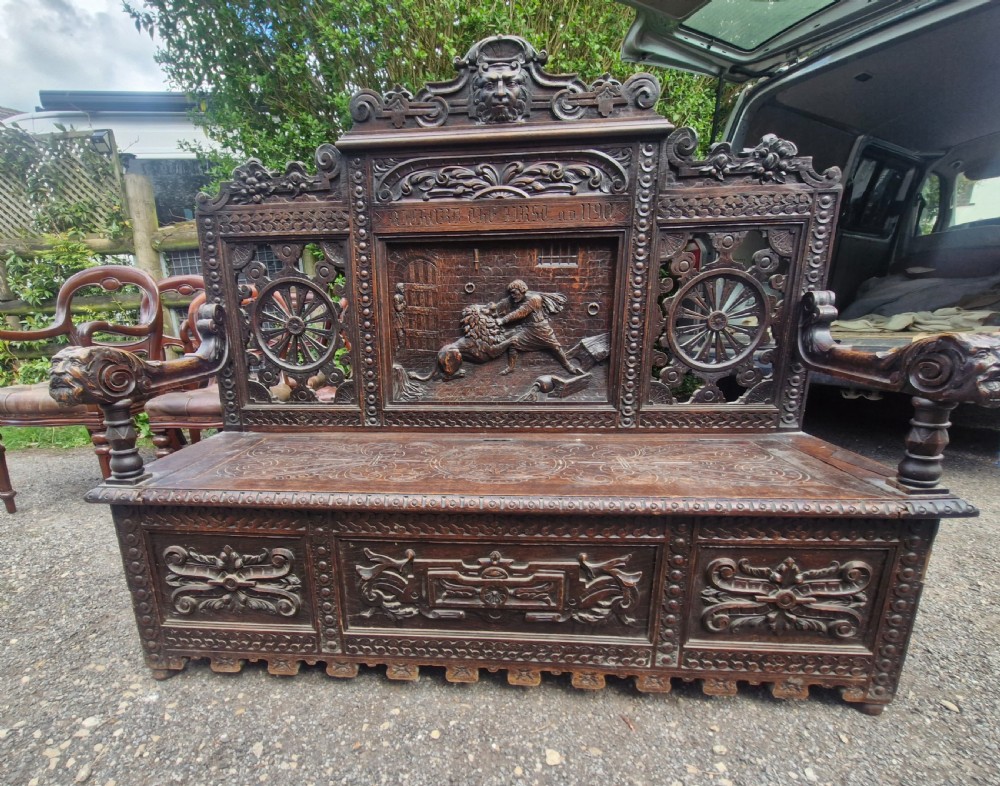 A late nineteenth-century dark carved oak hall bench featuring relief narrative carving, mask pediment, scroll arms, and a hinged seat. View II
