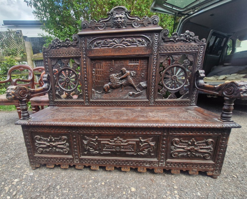A late nineteenth-century dark carved oak hall bench featuring relief narrative carving, mask pediment, scroll arms, and a hinged seat. View III