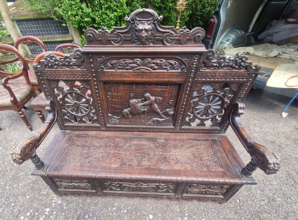 A late nineteenth-century dark carved oak hall bench featuring relief narrative carving, mask pediment, scroll arms, and a hinged seat. View VI