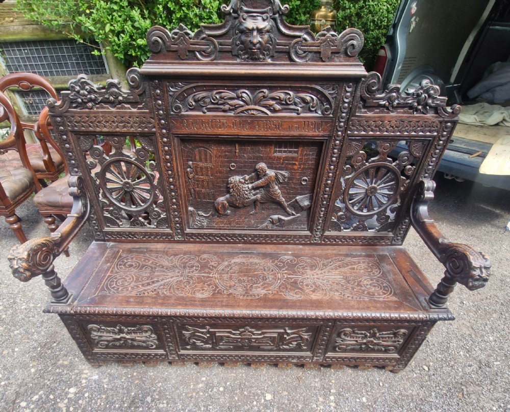 A late nineteenth-century dark carved oak hall bench featuring relief narrative carving, mask pediment, scroll arms, and a hinged seat. View VIII