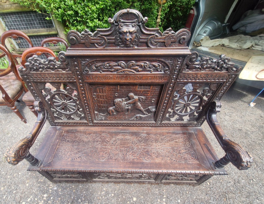 A late nineteenth-century dark carved oak hall bench featuring relief narrative carving, mask pediment, scroll arms, and a hinged seat. View IX