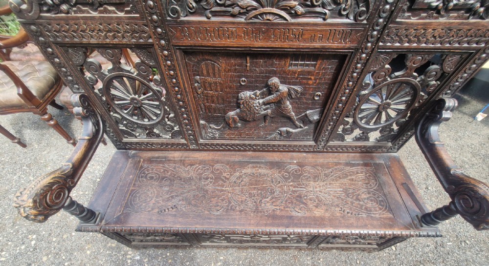 A late nineteenth-century dark carved oak hall bench featuring relief narrative carving, mask pediment, scroll arms, and a hinged seat. View X