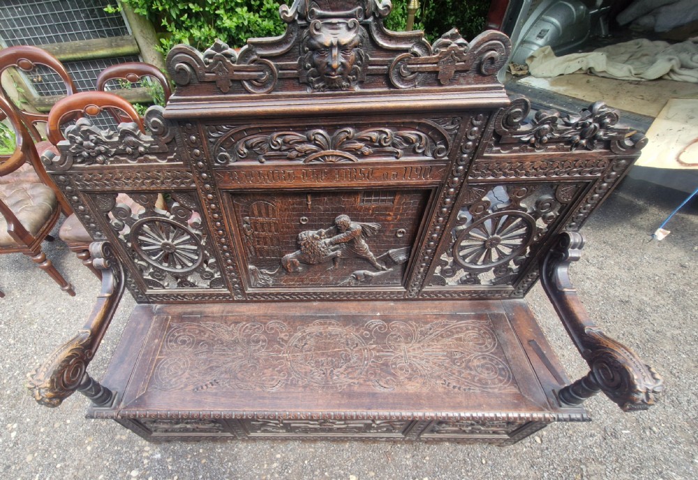A late nineteenth-century dark carved oak hall bench featuring relief narrative carving, mask pediment, scroll arms, and a hinged seat. View XI