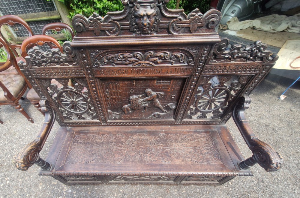 A late nineteenth-century dark carved oak hall bench featuring relief narrative carving, mask pediment, scroll arms, and a hinged seat. View XII
