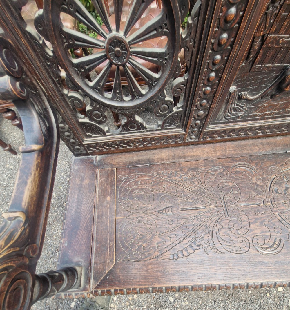 A late nineteenth-century dark carved oak hall bench featuring relief narrative carving, mask pediment, scroll arms, and a hinged seat. View XIII