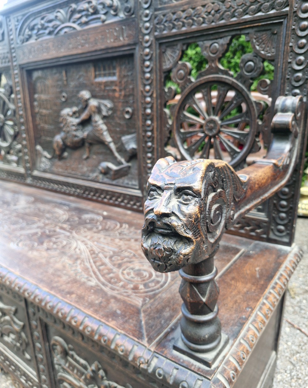 A late nineteenth-century dark carved oak hall bench featuring relief narrative carving, mask pediment, scroll arms, and a hinged seat. View XV