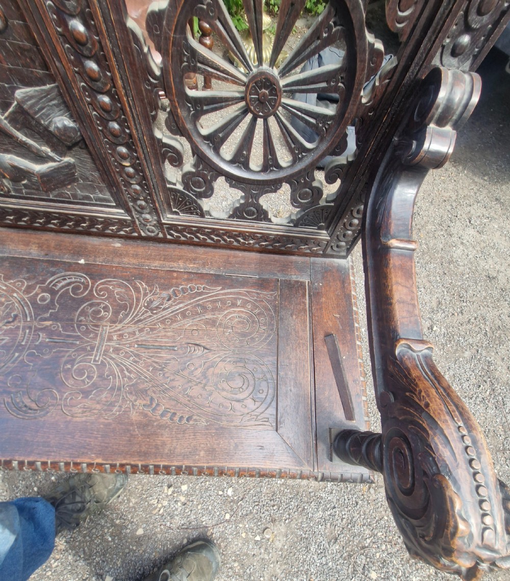 A late nineteenth-century dark carved oak hall bench featuring relief narrative carving, mask pediment, scroll arms, and a hinged seat. View XVII