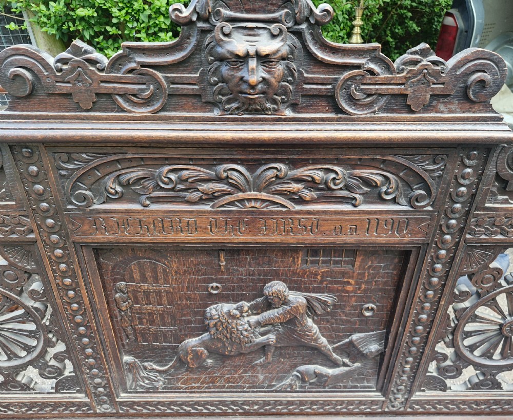 A late nineteenth-century dark carved oak hall bench featuring relief narrative carving, mask pediment, scroll arms, and a hinged seat. View XVIII